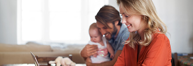 Shot of a young woman using a laptop with her husband and baby sitting in the background